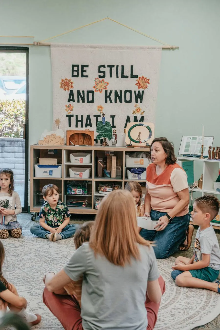 Children in the Godly Play room beneath a Be Still and Know banner.